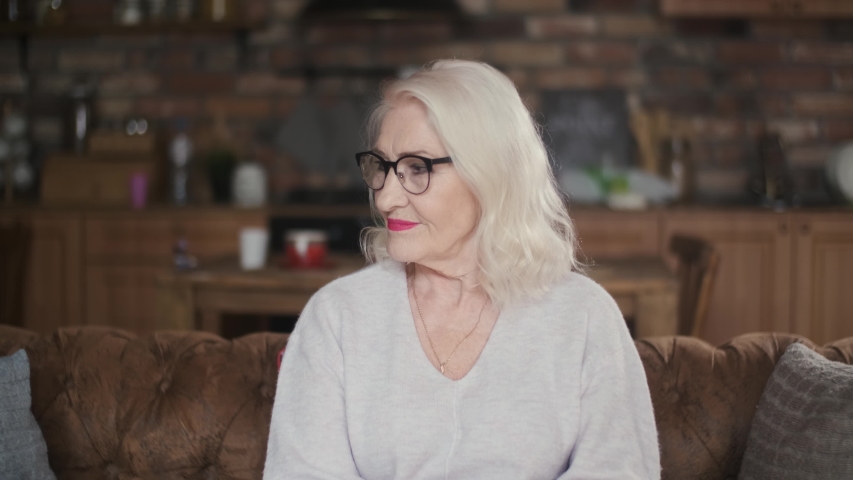 Elderly, beautiful woman with gray hair smiling looking at the camera. Portrait of a 70 year old grandmother with glasses. Happy senior citizen. An elderly, business woman sits on a sofa while being Royalty-Free Stock Footage #1054575896
