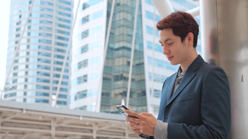 Happy overjoyed Asian Businessman holding phone celebrate good mobile news surprised at work with modern office building, excited man winner screaming yes rejoicing success looking at cellphone. | Shutterstock HD Video #1054583837
