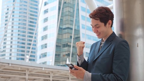 Happy overjoyed Asian Businessman holding phone celebrate good mobile news surprised at work with modern office building, excited man winner screaming yes rejoicing success looking at cellphone.