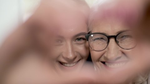 Happy elderly mom and daughter make a heart shape hand gesture. Look at camera bonding laughing showing family love concept, closeup portrait.