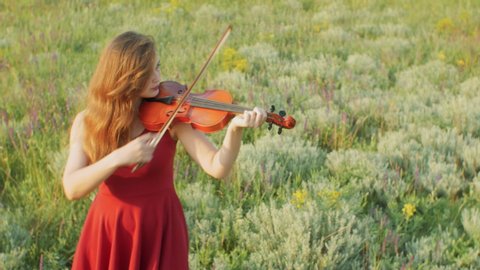 Beautiful girl violinist playing violin on summer meadow, young woman in red dress enjoying musical art walking on nature,shooting with a crane