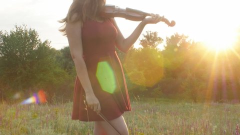 silhouette girl violinist playing violin on summer meadow, young woman in red dress enjoying musical art walking on nature at sunset