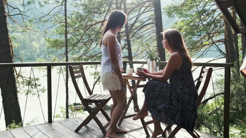 Two attractive girls are sitting at a coffee table on the balcony, enjoying nature. Two girlfriends drink tea outdoors on vacation.