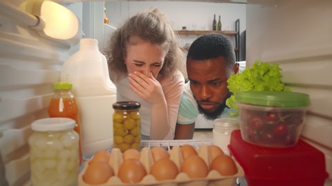 View from inside fridge of young multiracial couple feeling bad smell of spoiled meal. Diverse man and woman seeing awful smell of rotten food in refrigerator