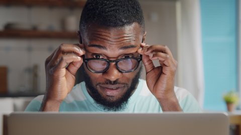 Tired young afro-american man in glasses with laptop computer working and rubbing his nose bridge at home office. Exhausted student massaging eyes studying at home