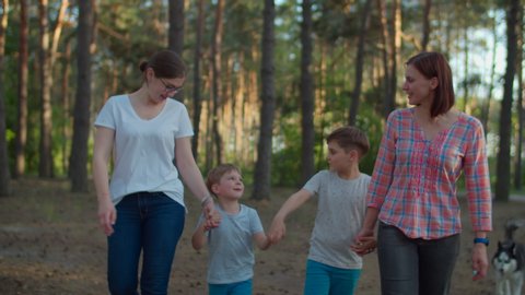Happy family of two moms and two sons talking and laughing while walking in summer forest with Siberian Husky dog. Slow motion, steadicam shot.