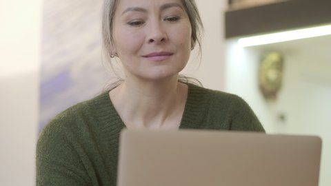 Smiling attractive mature woman with long gray hair turning off the laptop computer then looking away while sitting by the table indoors