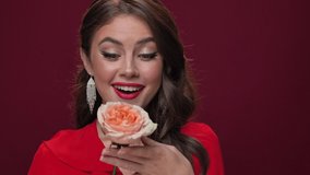 A close-up view of a happy young woman wearing a red dress is posing with a rose flower looking to the camera and rejoicing isolated over burgundy background