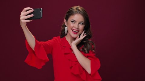 A fashionable attractive young woman wearing a red dress is taking selfie-photos using her smartphone isolated over burgundy background