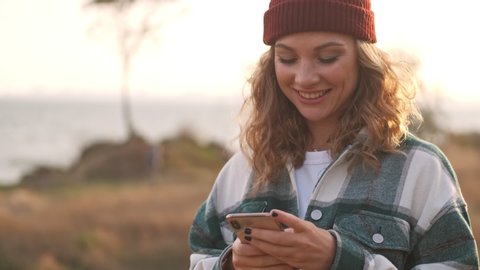 Smiling blonde woman wearing hat and plaid shirt using smartphone outdoors