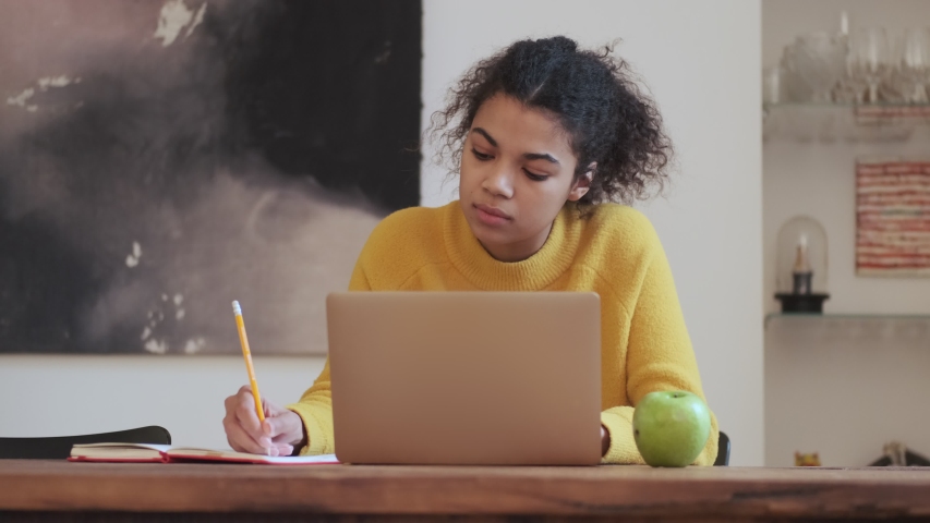 Calm african attractive woman writing something in notebook and using laptop computer while sitting by the table indoors