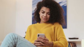 Smiling pensive african attractive woman using smartphone then looking away while sitting on chair indoors