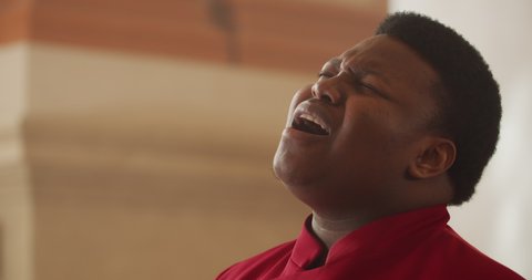 Side view of male performer wearing in red stylish suit singing emotionally. Portrait of afro american man performing worship music in church. Concept of religion. Blurred background.