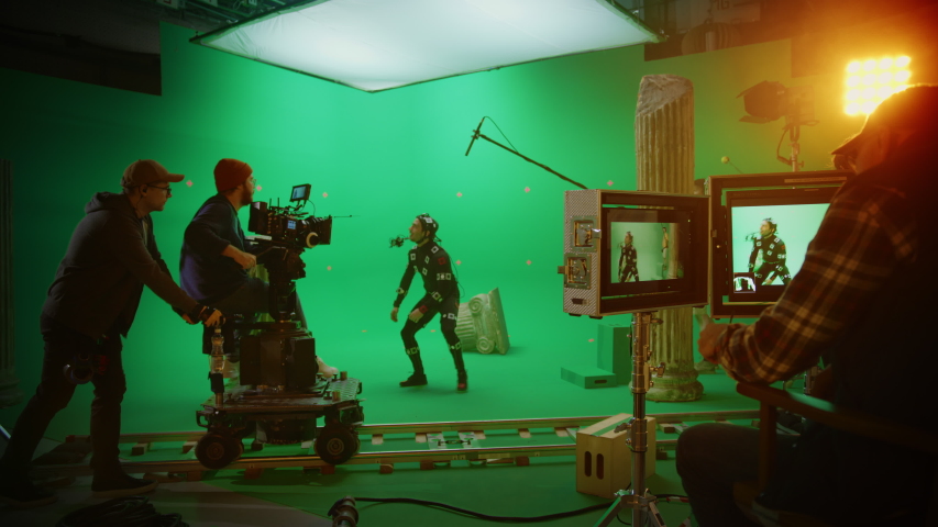 In the Big Film Studio Professional Crew Shooting Blockbuster Movie. Director Commands Cameraman to Start shooting Green Screen CGI Scene with Actor Wearing Motion Tracking Suit and Head Rig Royalty-Free Stock Footage #1054601096
