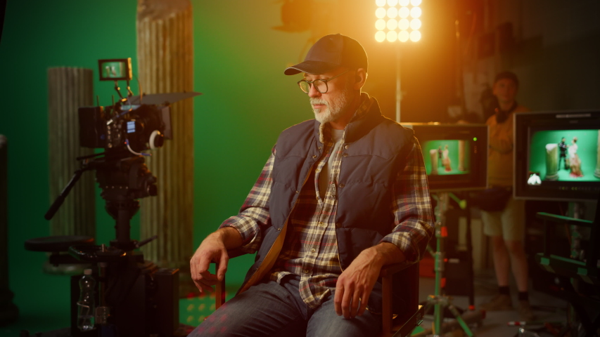 Protrait of Prominent Successful Senior Director Sitting in a Chair on a Break Looking at Camera Cheerfully. On the Studio Film Set with High End Equipment Professional Crew Shooting High Budget Movie