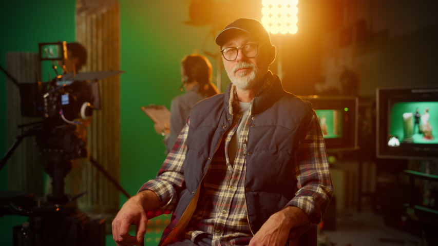 Protrait of Prominent Successful Senior Director Sitting in a Chair on a Break Looking at Camera Cheerfully. On the Studio Film Set with High End Equipment Professional Crew Shooting High Budget Movie
