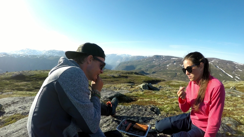 A couple in hiking outfit camping in the wilderness, at the top of fjord with the view on Eidfjord. They are preparing hot dogs from the grill and sipping a beer. Being close to nature. Having fun
