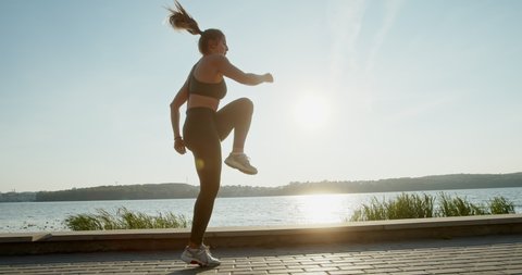 Tracking shot of slim sportswoman jumping forward and running on spot during fitness training on lake embankment during sunset