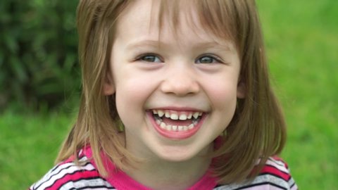 Portrait of little girl in summer park sits on green lawn looks at camera, smiles and laughs. Close up. Outdoors. Happy сhild plays in nature