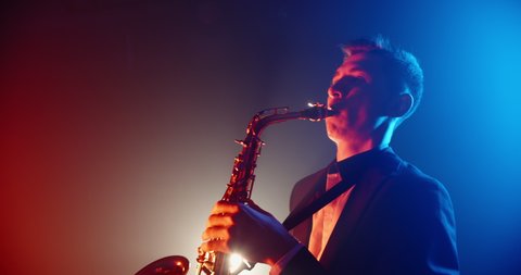 Saxophonist wearing a suit performing a solo on stage with band, spotted by red and blue lights of club - music, art concept 4k footage