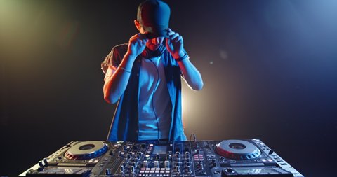 Cool hipster disc jockey performing in a nightclub at a mixer controller, spotted by colorful lights on smoked black background - nightlife concept 4k footage