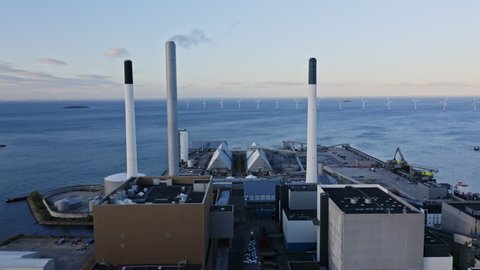 Drone shot of industrial buildings and a view of their rooftop, with long chimneys emitting white steam, as the drone tracks forward and shows a beautiful view of the ocean waters and windmills