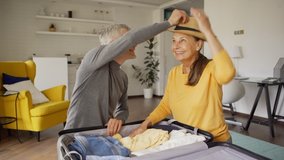 Tilt up medium shot of excited senior couple talking cheerfully while packing suitcase for vacations together. Joyful woman trying on straw hat. Happy couple closing bag and embracing - Powered by Shutterstock - Get 15% off with code: PIKWIZARD15