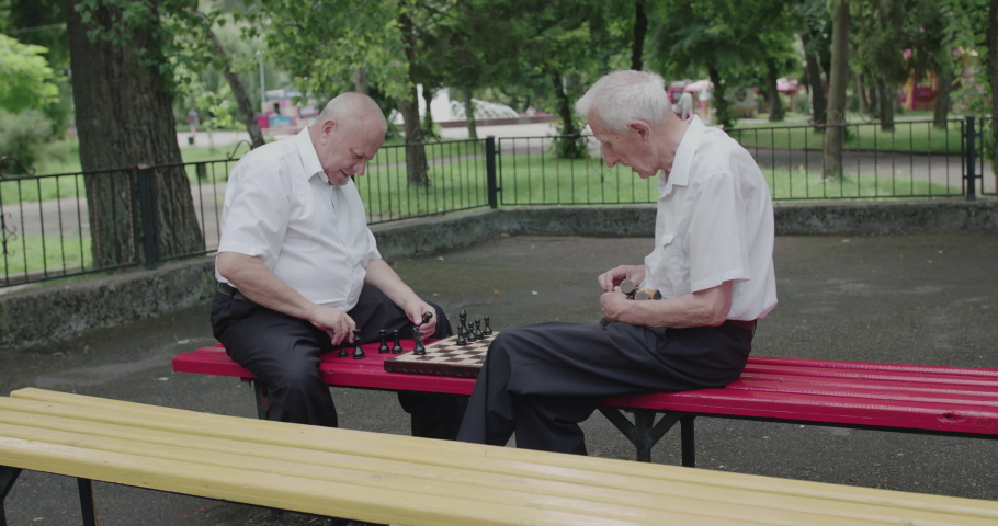 Two seniors friends playing the chess on street bench on leisure at summertime. 4K