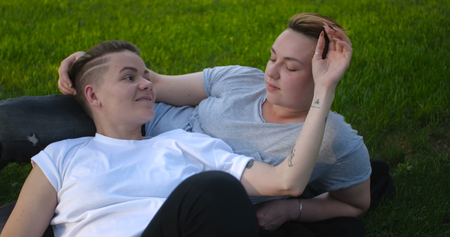 Close up portrait of female same-sex couple lying on the grass. Two pretty stylish girls with short creative hairstyles hug and kiss to hand each other in love. 4k slow motion 50 fps