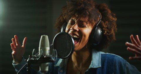 An young professional african female singer is performing a new song with a microphone while recording it in a music studio. 