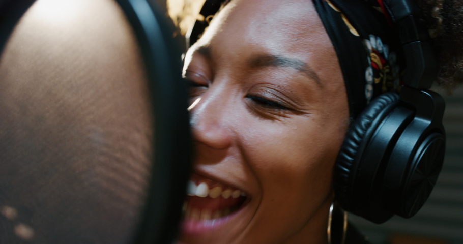 Close up of an young professional smiling energetic african female singer wearing bandana and headphones is performing a new song with a microphone while recording it in a music studio.  Royalty-Free Stock Footage #1054633787