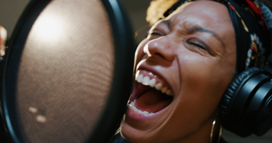 Close up of an young professional smiling energetic african female singer wearing bandana and headphones is performing a new song with a microphone while recording it in a music studio. 