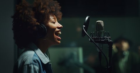 A professional male sound producer with headphones is recording a new song with an young african female singer in a music studio.
