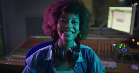 An young happy professional african female sound producer is taking off headphones while recording a new song and smiling satisfied in camera in a music studio with colorful lights on a background.