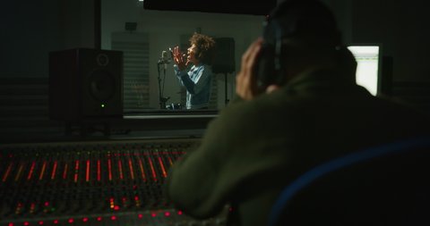 Back view of professional male producer with headphones is recording a new song with young african female singer in a music studio.
