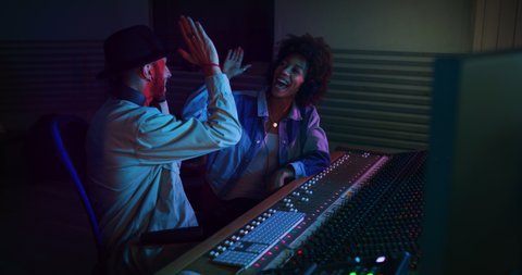 A professional male producer and young african female singer are happy with results of recording a new song and  are celebrating a future success in a music studio with colorful lights on a background