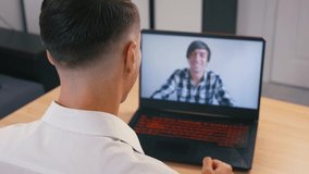 Online conference. A business man greets his work colleague and communicates with him remotely from home by webcam. Video call from a laptop. Over shoulder close up view