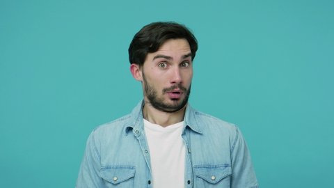 Closeup of surprised astonished bearded guy in jeans shirt standing, staring at camera with amazement, keeping mouth open, shocked by unexpected news. indoor studio shot isolated on blue background