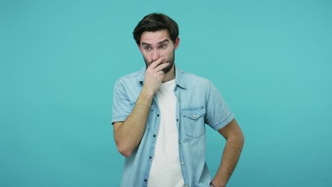 No, I won't say anything. Shocked confused scared bearded guy in jeans shirt covering mouth with hand, looking depressed and amazed by horrible truth secret. studio shot isolated on blue background