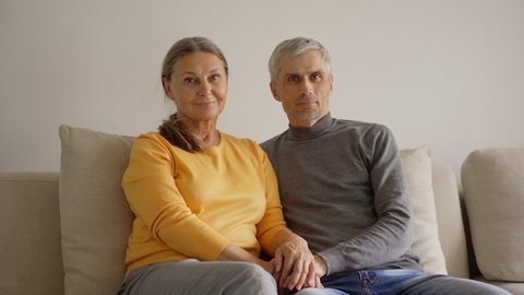 Panning medium shot portrait of beautiful and loving senior couple looking at camera sitting on sofa at home and holding hands