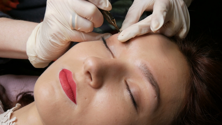 Beautician in black gloves makes permanent makeup correction of the shape of the eyebrows to a young beautiful girl. Microblading, eyebrow tattoo in a beauty salon. | Shutterstock HD Video #1054644293