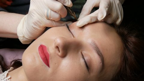 Beautician in black gloves makes permanent makeup correction of the shape of the eyebrows to a young beautiful girl. Microblading, eyebrow tattoo in a beauty salon.