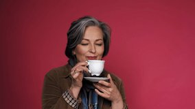 Charming mature woman enjoys cup of coffee with eyes closed. Happy gray-haired lady drinks coffee posing on pink background in the studio. Close up portrait with text space on right. Prores 422. 