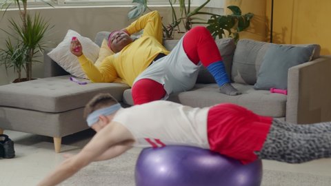 Humor in sports. Fat african american lazy guy resting on couch while his clumsy funny friend doing fake gymnastics with fitness ball.