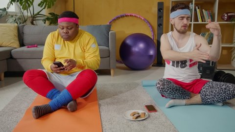 Comical scene. Black clumsy guy eating cookies taking smartphone pictures of his concentrated funny friend meditating. Yoga at home. Sports parody.