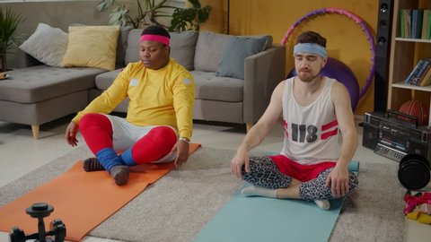 Fat african american man doing yoga with bearded fitness instructor, sitting lotus pose on mats, doing meditating moves for deep breathing. Training. Fun concept.