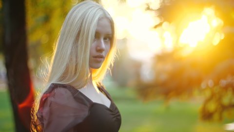 Attractive young scandinavian blonde teen girl walking in summer sunset city park, looks at camera with alluring look. Confidence fashion norwegian stylish teenager walking in sunlight shining