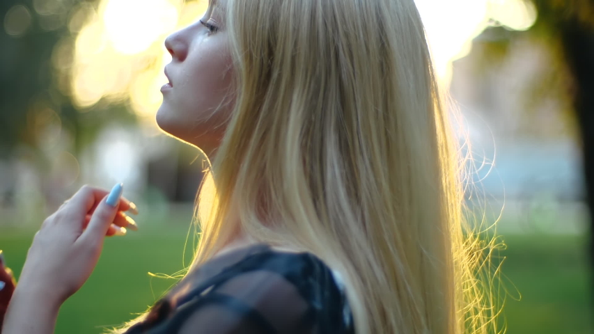 Close-up profile stunning young blonde in lightweight dress fluffy light long  blonde hair in park in sunshine. Beautiful girl enjoys leisure time. Сare for natural colored delicate blond hair Royalty-Free Stock Footage #1054651496