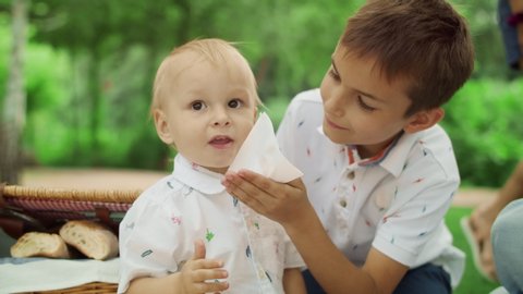 Portrait of elder brother taking care about small boy at picnic. Cheerful boy wiping toddler face with napkin. Mother hand giving cherry to little boy. Happy family spending time outdoors