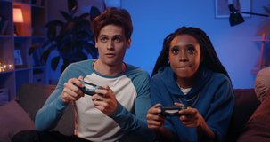 Happy boyfriend and girlfriend playing video games and holding joysticks at home. Millennial couple enjoing game while sitting on sofa and spending free time. Concept of relationship.
